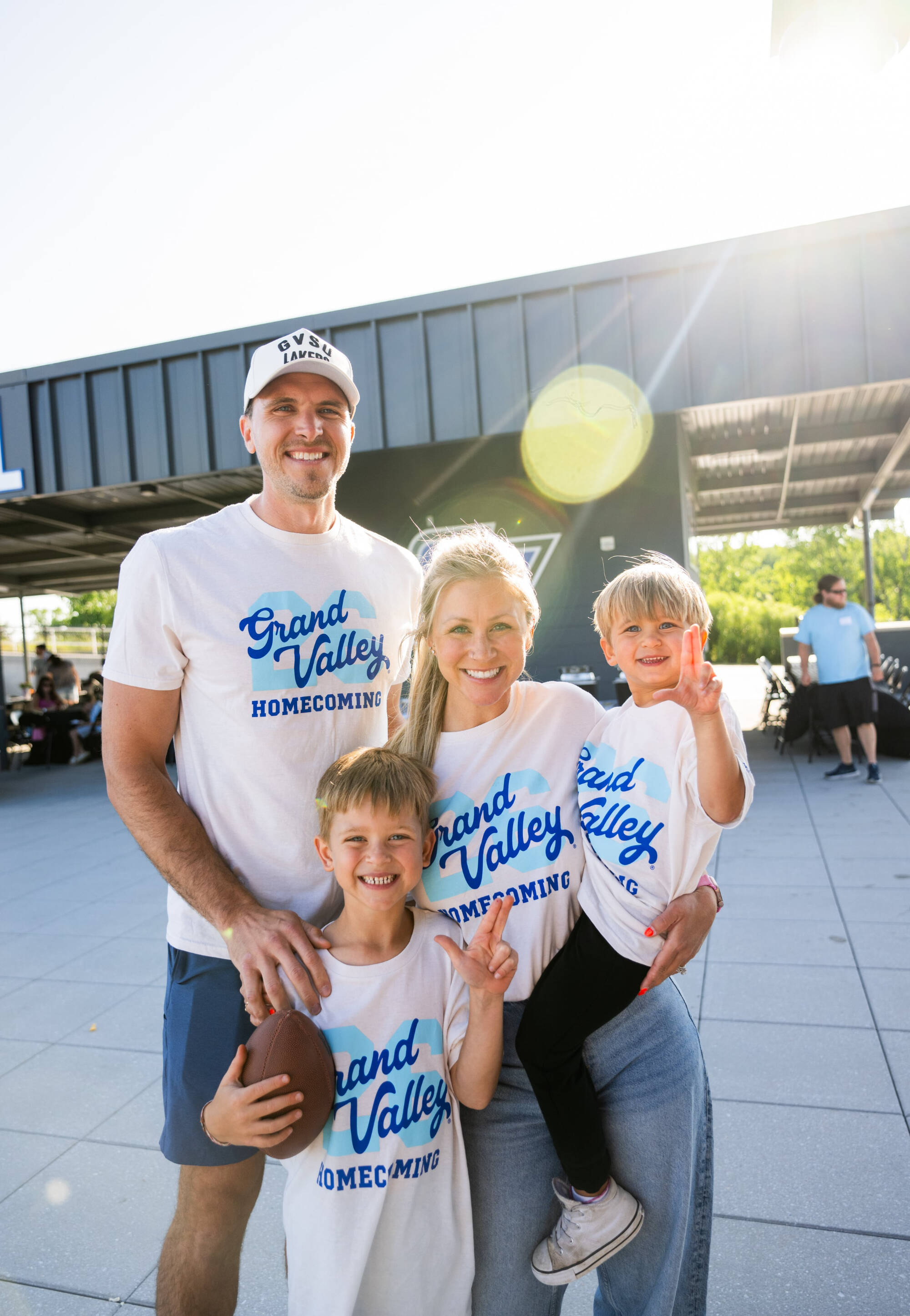 Laker Family smiles wearing GVSU Homecoming t-shirts in Lubbers Stadium with sun shining behind